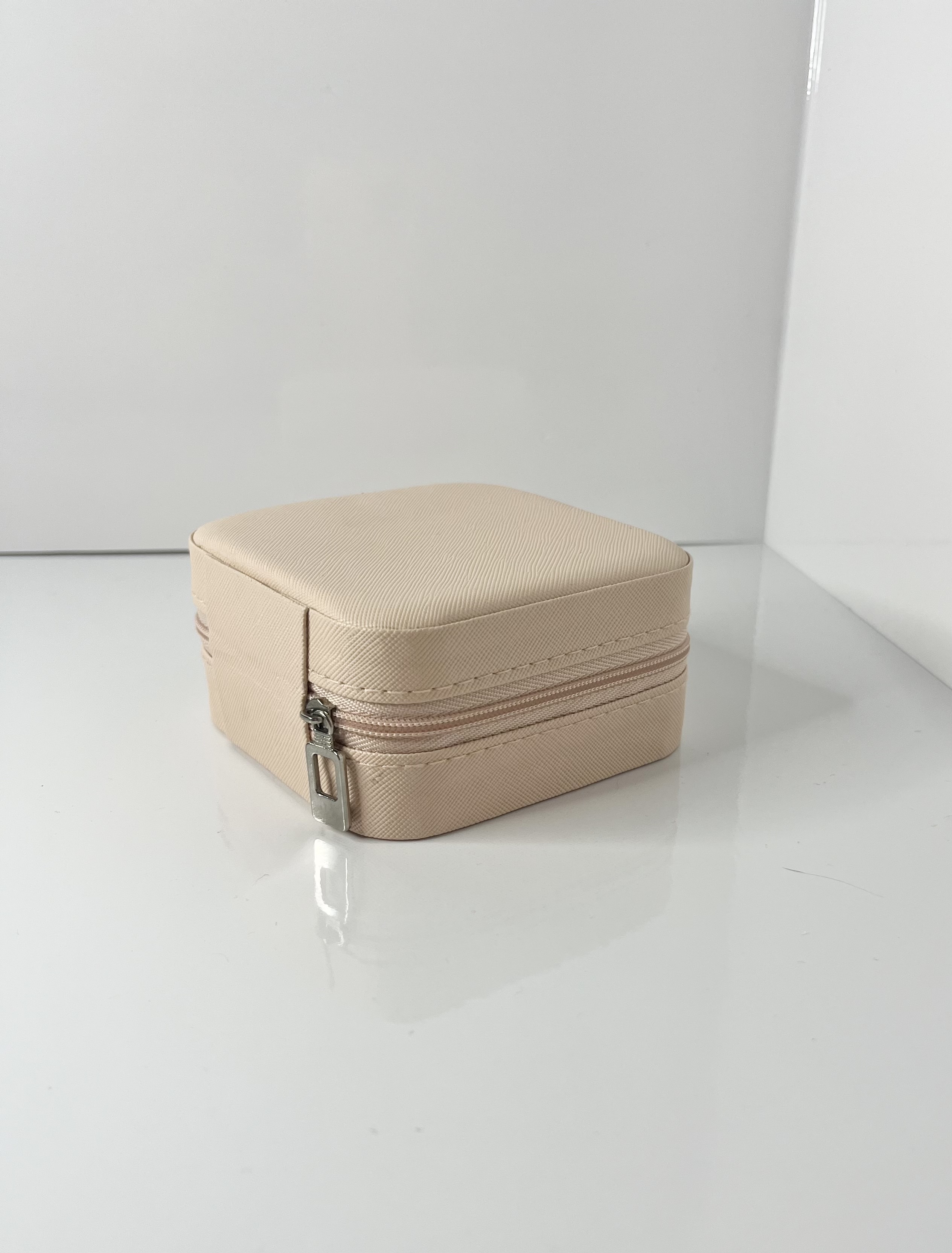 A beige jewelry box is shown from a diagonal angle, highlighting its cube form and zipper detail.