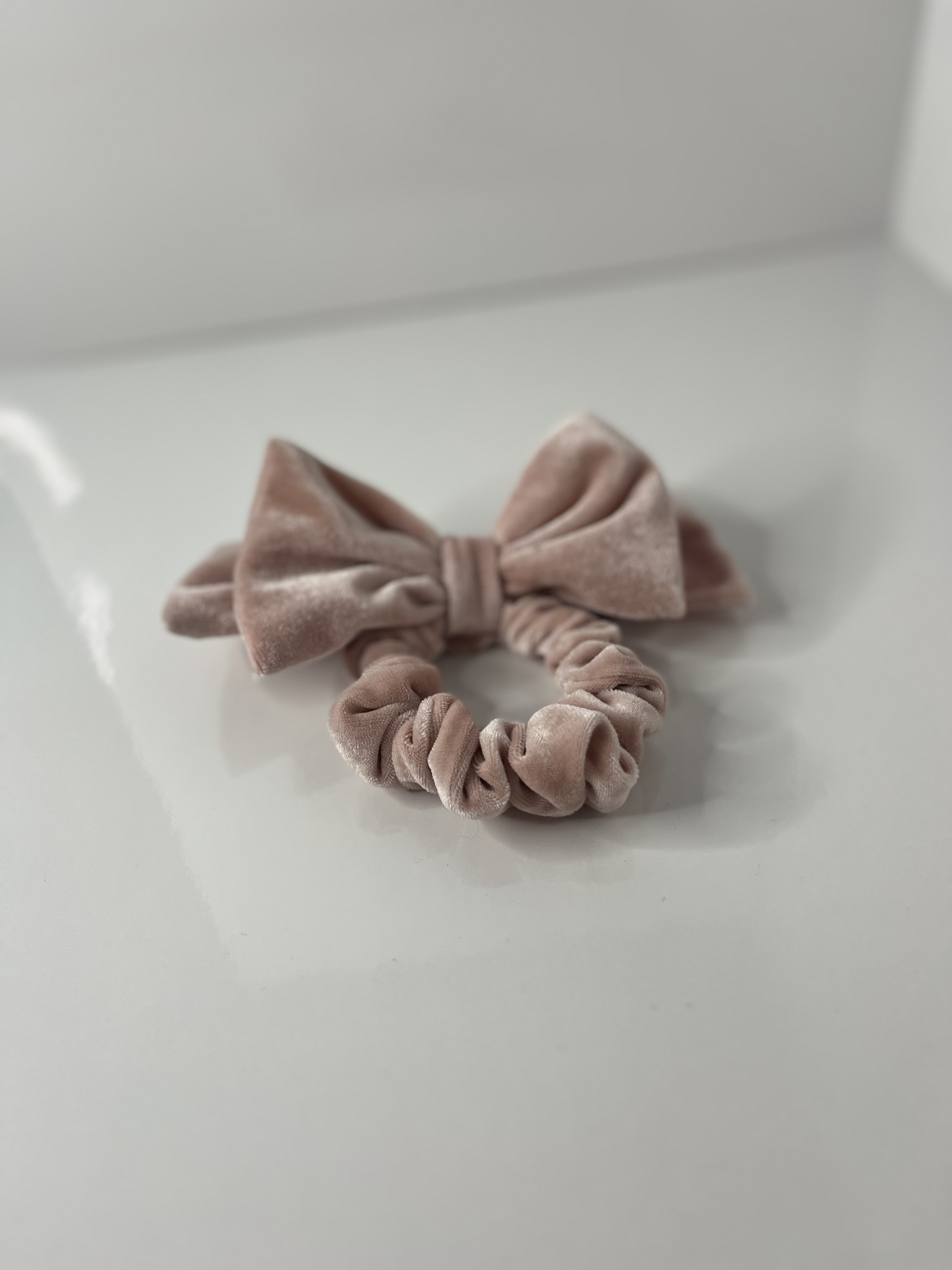 A pink velvet scrunchie with an attached bow rests on a flat surface, its torus shape loosely defined by gathered folds.