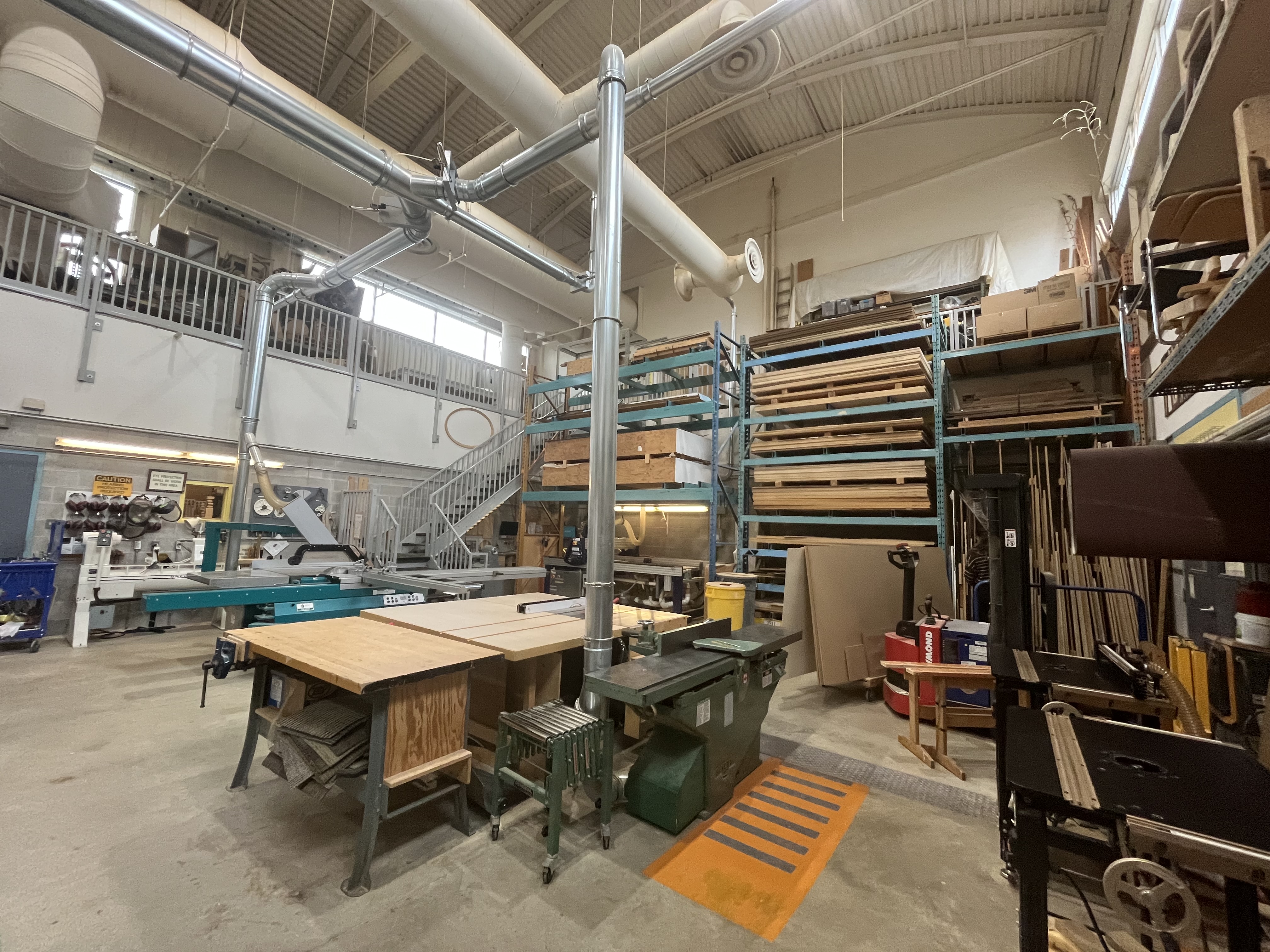 The woodshop contains industrial-grade cutting tools, organized lumber storage, and a central workbench for hands-on fabrication.
