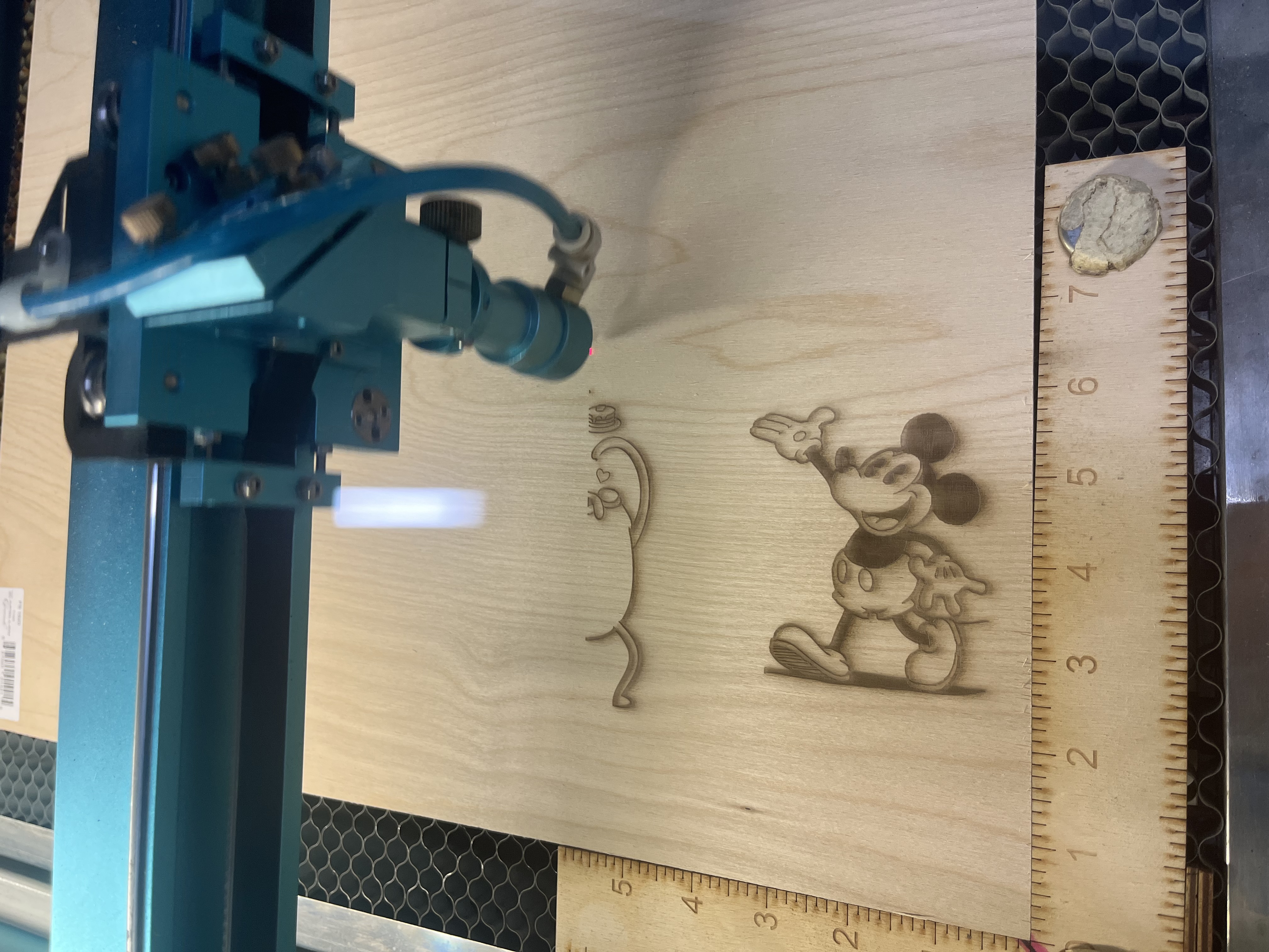 The machine engraves various character designs and logo elements onto the wooden surface, leaving precise recessed burn lines.