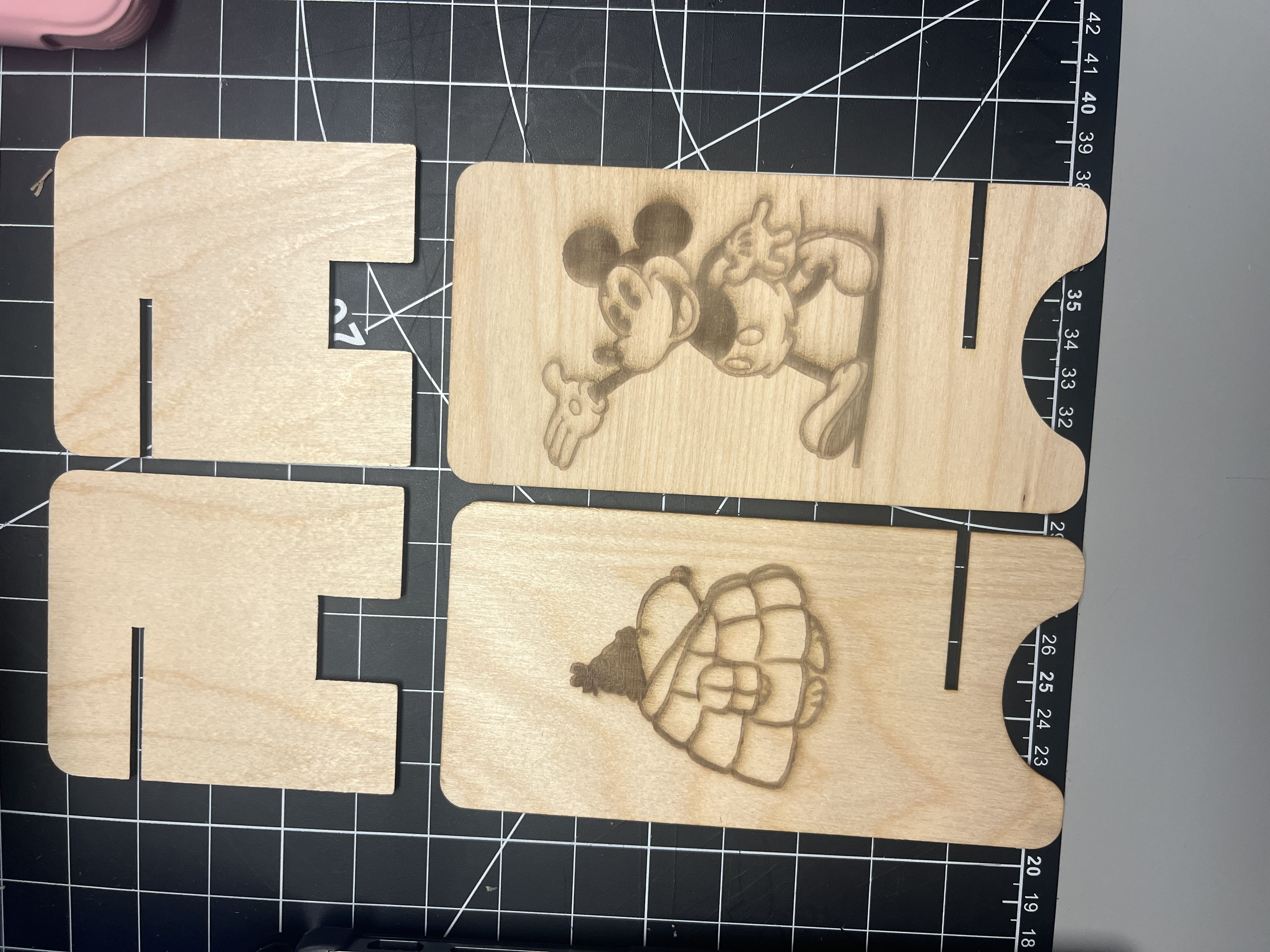 Four individual laser-cut plywood phone-stand components are displayed, including two structural base pieces and two engraved decorative panels featuring illustrated characters.